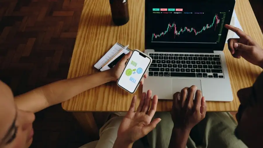 A laptop and a phone on a table, one showing a chart and the other a buy-and-sell screen